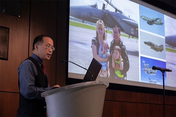 man at podium wearing glasses and vest shows slides behind him of military service