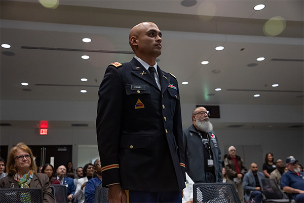 man in military uniform stands in foreground of audience where other veterans stand for recognition