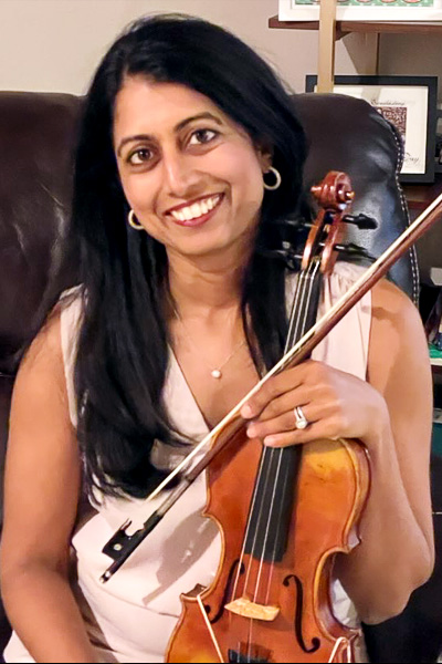 Smiling woman with long dark hair wearing a white dress, seated in a dark leather chair, holding her violin. 