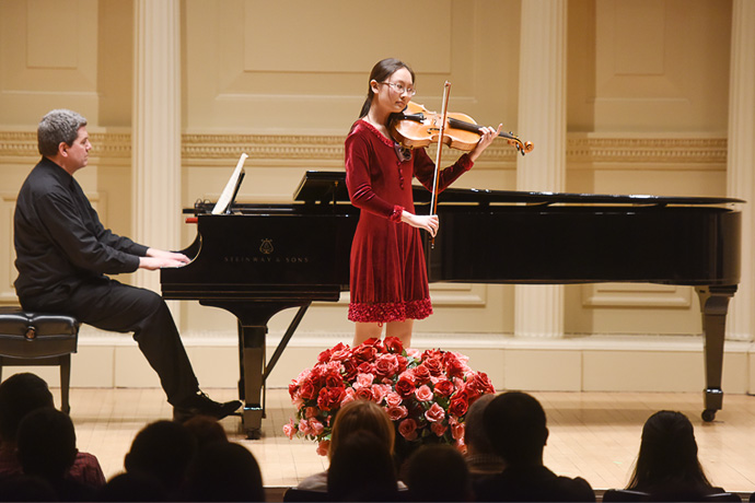 Woman with long dark hair wearing a red dress, standing next to a grand piano, playing her violin. A man with dark hair and dark clothing is playing the piano.