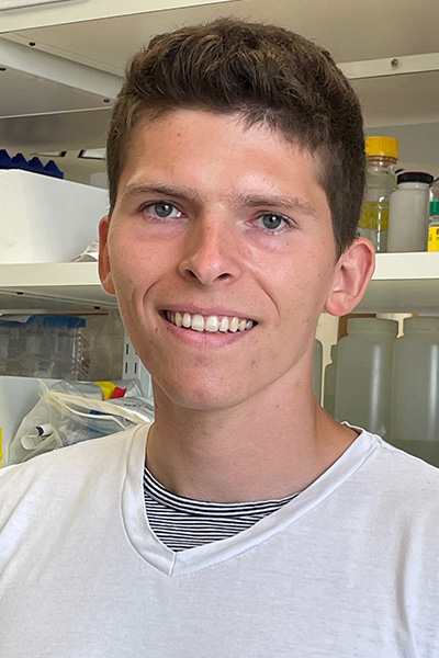 Smiling man with short dark hair wearing a white tee shirt, standing in a lab.