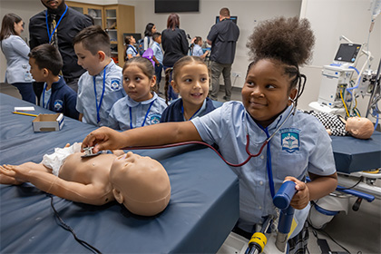 group of 5 kids watch as little girl grins while using a stethascope on a baby medical dummy