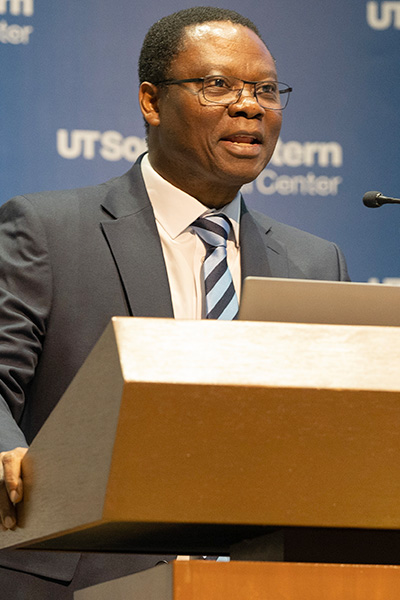 A man with dark hair, wearing glasses and a gray suit lectures from a podium. Blue background says UTSouthwestern Medical Center