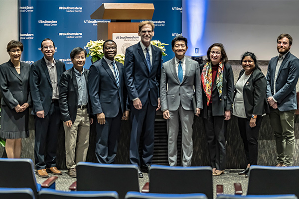 A group of 6 men and 3 women stand in front of a stage, smiling for the camera.