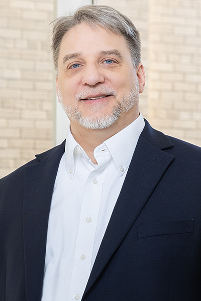 Smiling man with gray hair and beard, wearing a dark jacket over a white shirt.