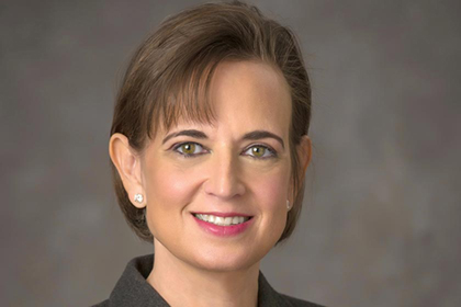 Smiling woman with short brown hair, wearing a lab coat and sitting in front of a screen showing micro-organisms.