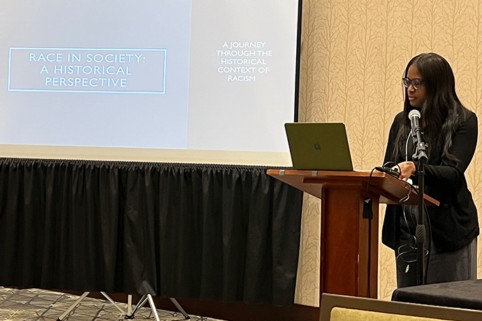 Woman with long dark hair, wearing glasses, speaking at a podium. On screen nearby, RACE IN SOCIETY: A HISTORICAL PERSPECTIVE - A JOURNEY THROUGH THE HISTORICAL CONTEXT OF RACISM.