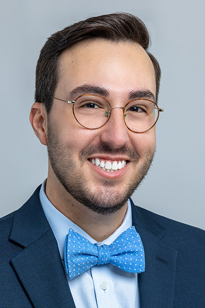 Smiling man with dark hair, trim beard, and mustache, wearing a gray suit, blue ruffled shirt, blue bow tie, and glasses.