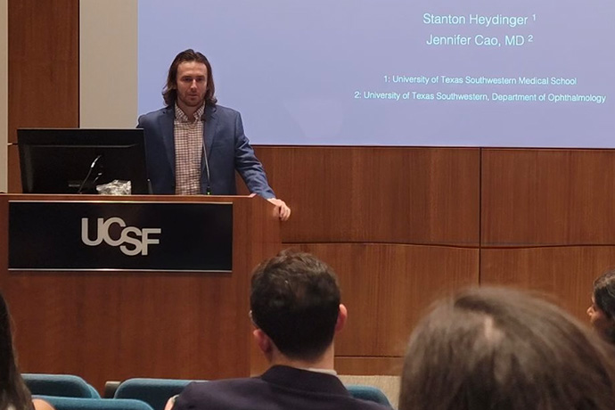 Man with brown hair and beard, standing at a podium facing an audience, in front of a screen with: Stanton Heydinger, University of Texas Southwestern Medical School, Jennifer Cao, MD, Univeristy of Texas Southwestern, Department of Ophthalmology