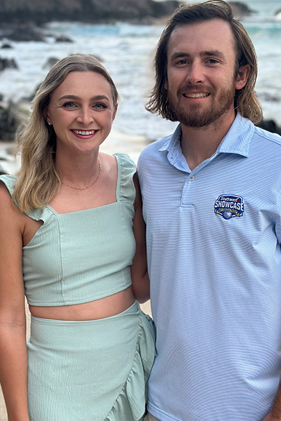 Smiling woman with ling blond hair, standing on a beach with smiling man wit brown hair, beard, and mustache.