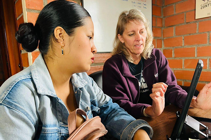 Two casually dressed woman sit a a desk looking at a computer monitor in a room with red brick walls.