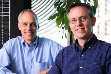 at left in back, white balding male wearing light blue shirt; in foreground right, white male wearing glasses and dark blue shirt