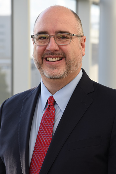 Smiling man with receding hair and graying beard wearing a dark suit with a light shirt and red tie.