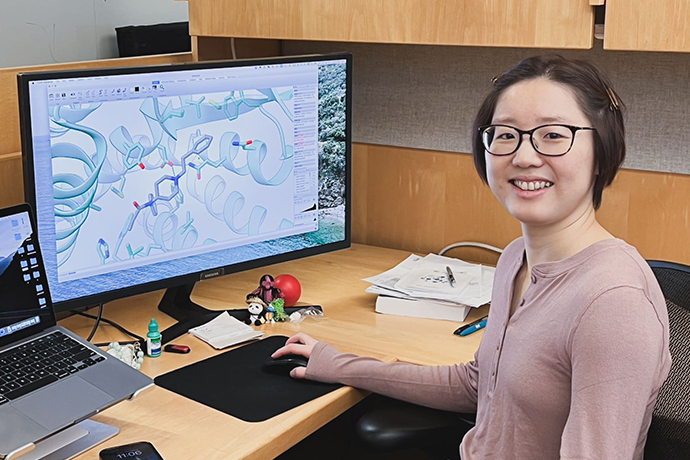 young Asian researcher with short dark hair and glasses wearing brown shirt sits at desktop computer showing graphic display of cellular research on monitor