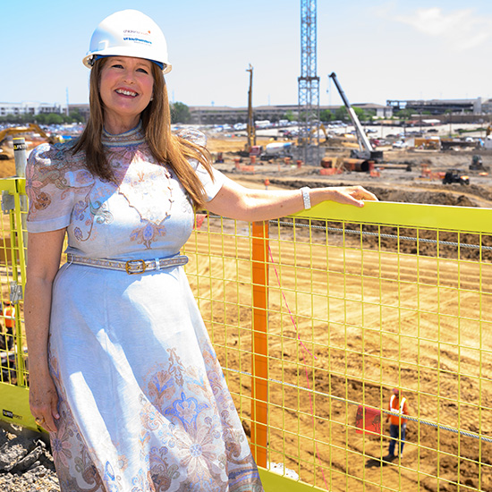 Woman wearing a hard-hat stands next to fence at construction site.