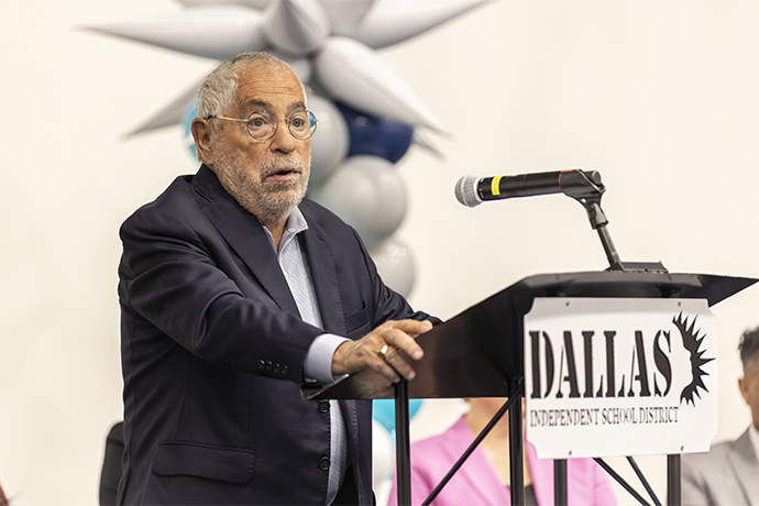 older man with round glasses, grey hair and beard speaks from podium