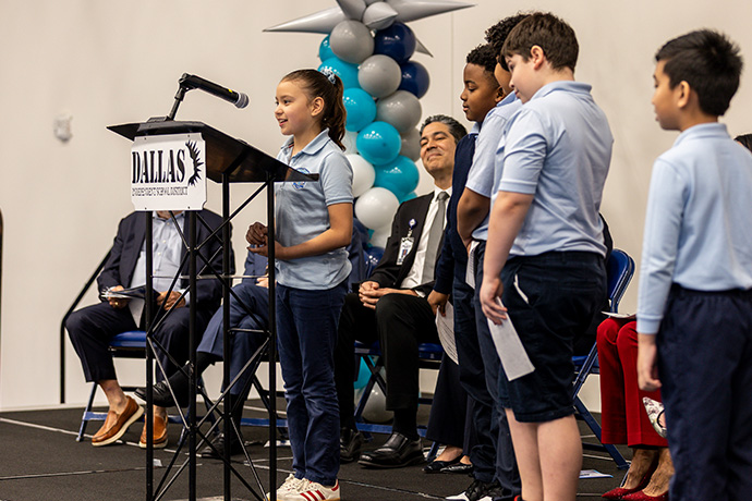5 young kids line up to speak with little girl standing behind podium as seated adults smile in the background