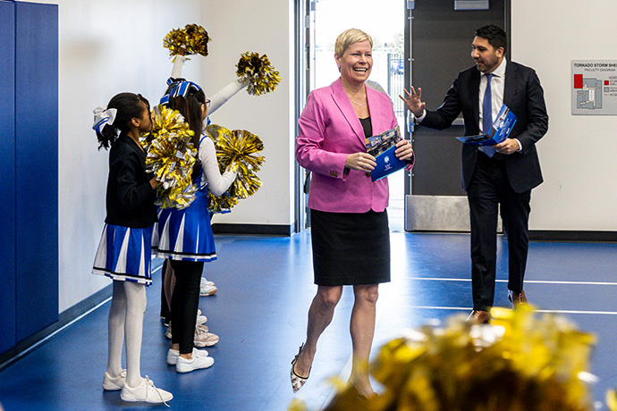 5 little girl cheerleaders wave gold pompoms as woman in pink jacket and man in dark suit enter room