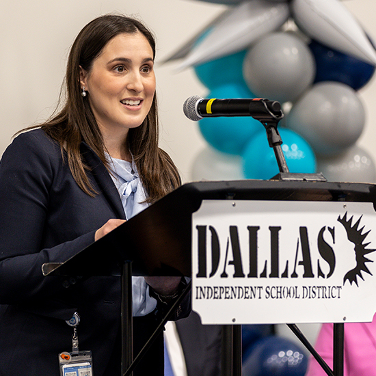 woman with long dark hair in black suit speaks from DISD podium