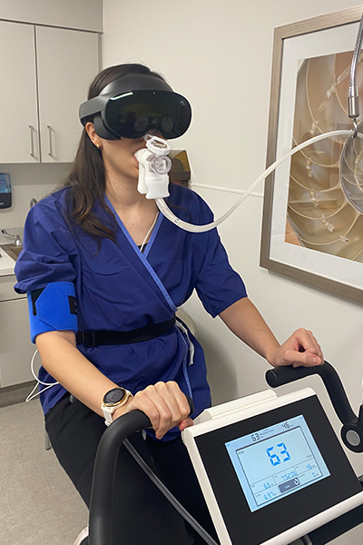 female in blue scrubs sits on exercise bike in lab wearing VR goggles and breathing into oxygen tube