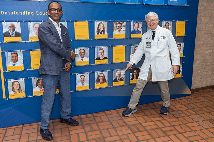 black man in blue suit with glasses and white-haired man in lab coat pose in front of honoree photos on wall