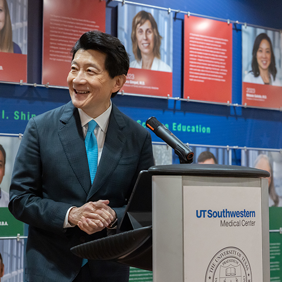 dark haired man in blue suit speaks from podium in front of wall of photos