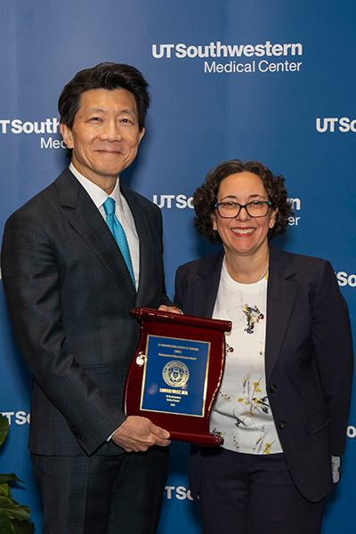 Smiling man in blue suit holding a plaque stands with woman in front of wall with pictues
