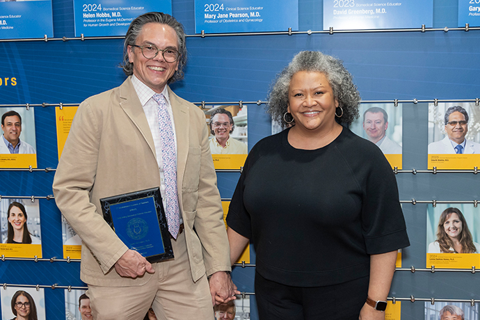 Smiling man holding a plaque stands with woman in front of wall with pictues