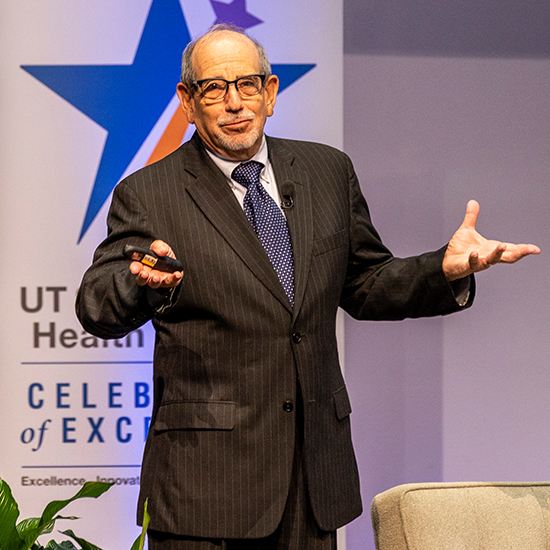 balding man with glasses in dark pinstripe suit gestures from CoE stage