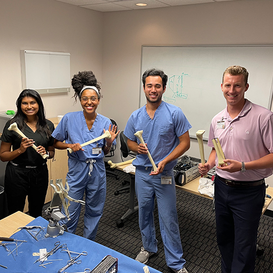 from left: short dark-haired woman in black, taller woman in blue scrubs, man in blue scrubs, and blond man in pink shirt; all smiling and holding model bones in classroom with clamps and equipment on table in foreground