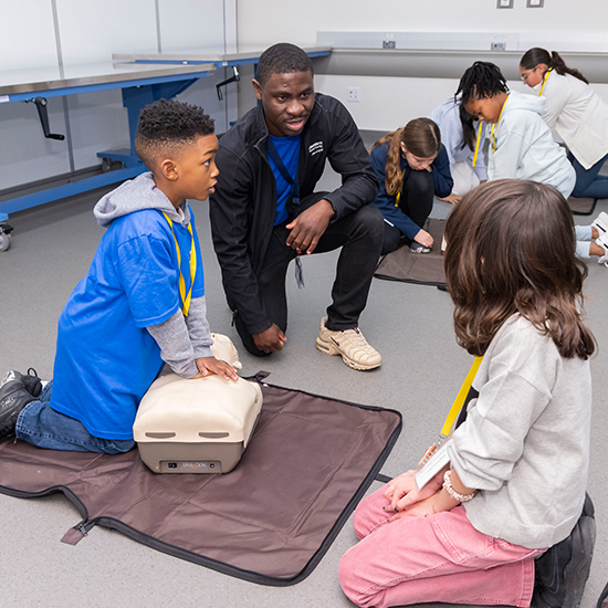 young man in black tracksuit knees next to mat where young boy in hoodie practices CPR on medical dummy; little girl watches as 4 other children practice on mats in background
