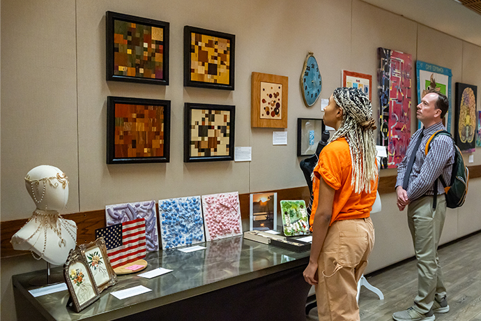 woman in orange shirt with long blonde braids looks at artwork on gallery wall; display table with more art pieces in front of her