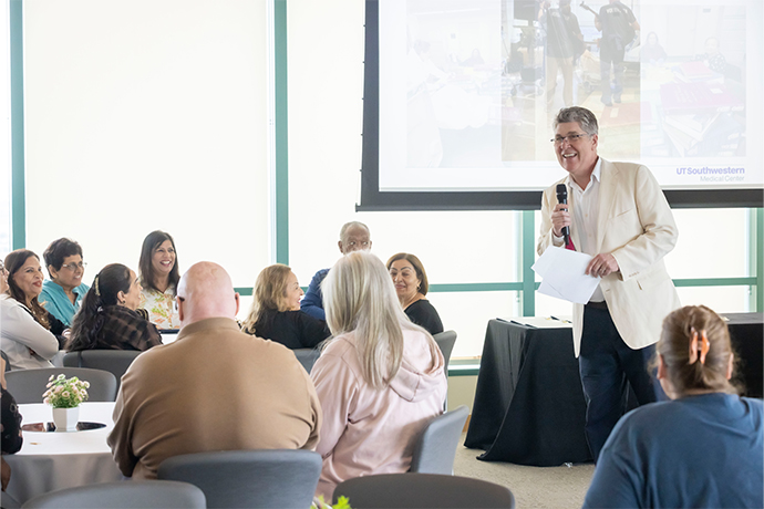 Smiling man speaks to an audience seated at round tables.