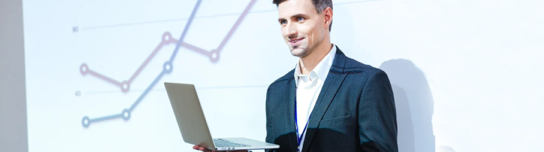 man in suit holds laptop and stands in front of wall projecting image of bar chart