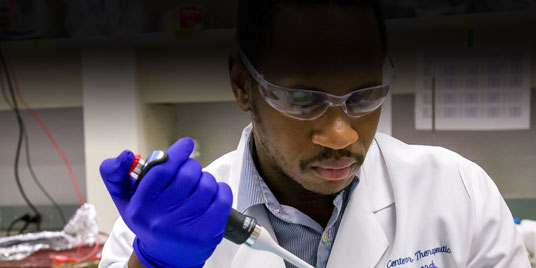 A male graduate student wearing a white lab coat, goggles, and blue gloves fills a test tube from a pipette