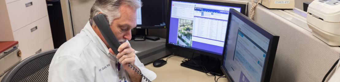 A gray-haired man sits at a desk with two computer monitors in front of him and holding a telephone to his ear