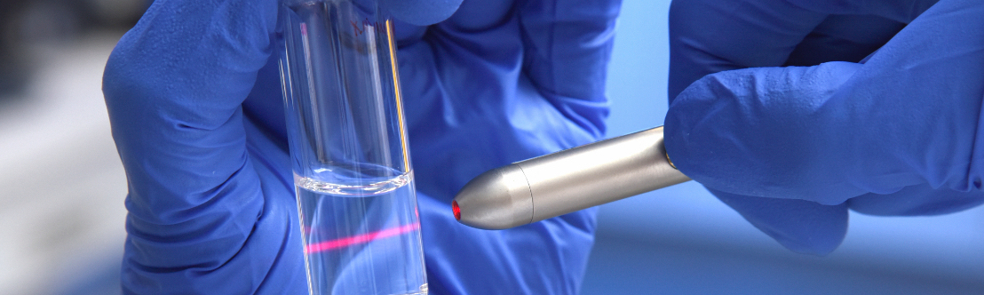 Held in blue gloved hands, a silver device shines a red light beam through a test tube filled with liquid