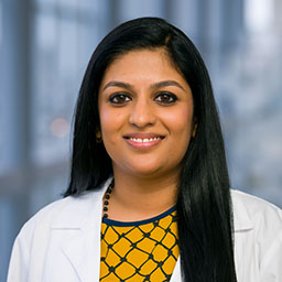 woman with long, dark hair wearing a gold and black patterned shirt and white coat