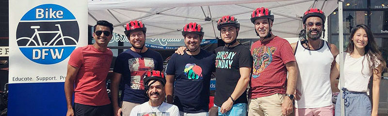 A line of men and women, wearing bike helmets next to an outdoor banner saying Bike DFW