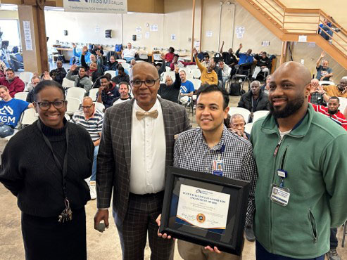 four people standing infront of a crowded room, one holding a framed certificate