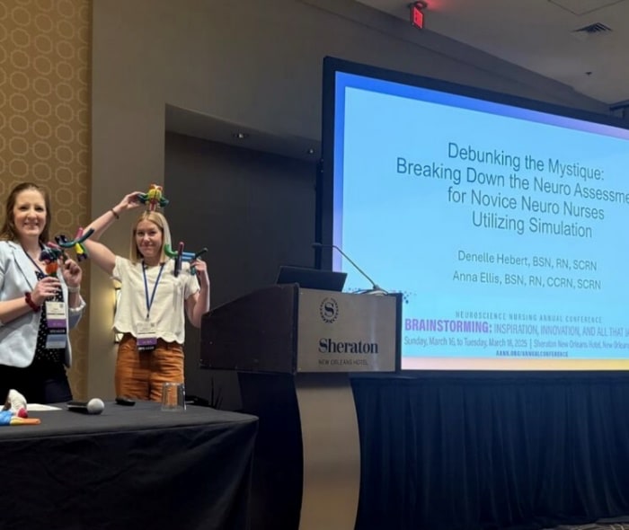 Two women with colorful props beside a screen that reads Debunking the Mystique: Breaking Down the Neuro Assessment for Novice Neuro Nurses Utilizing Simulation