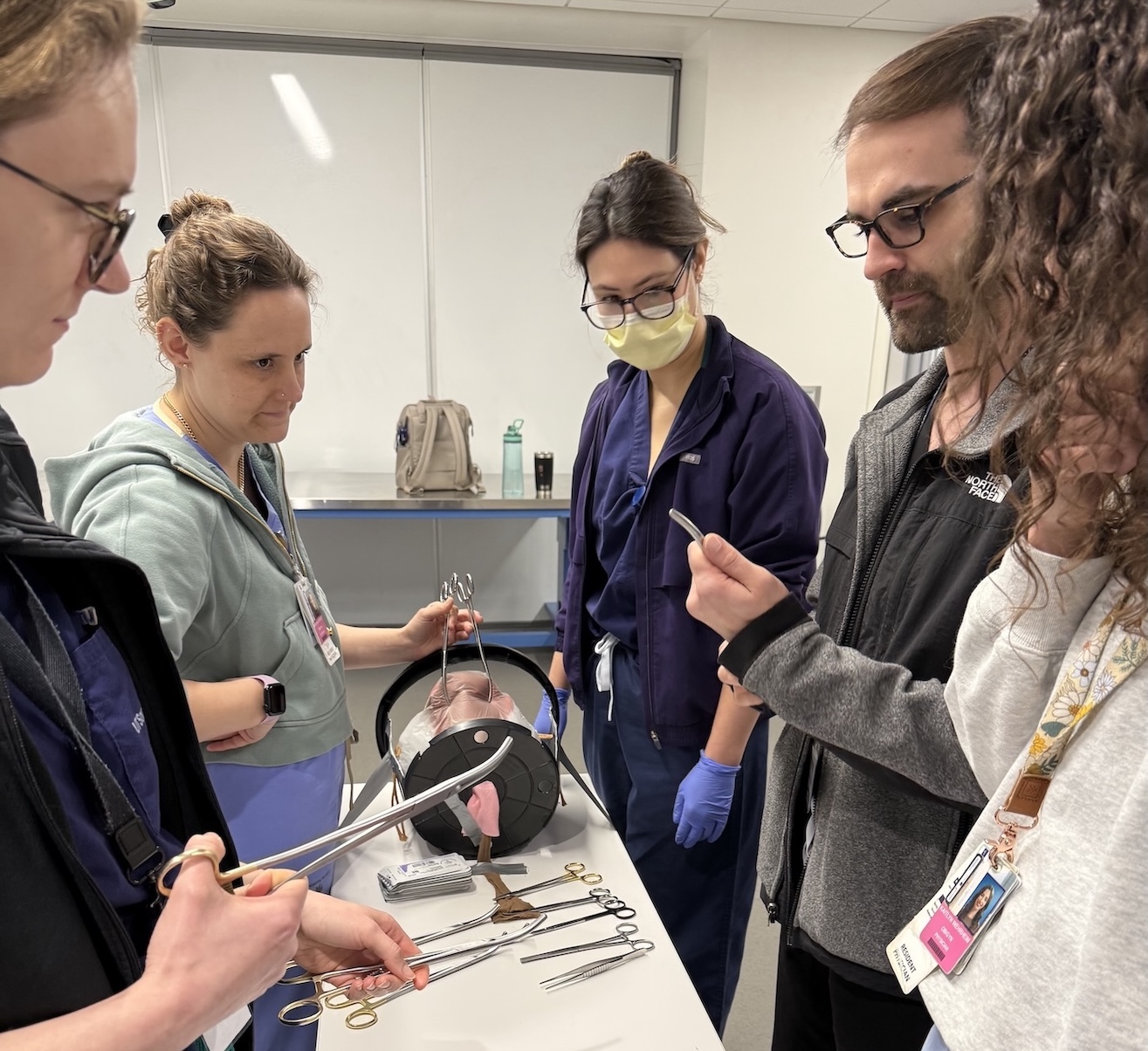 A small group of residents examines surgical instruments and simulation models at a workstation.