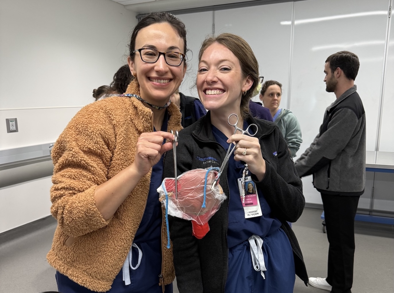 Two residents hold surgical instruments and a practice model during a lab session.