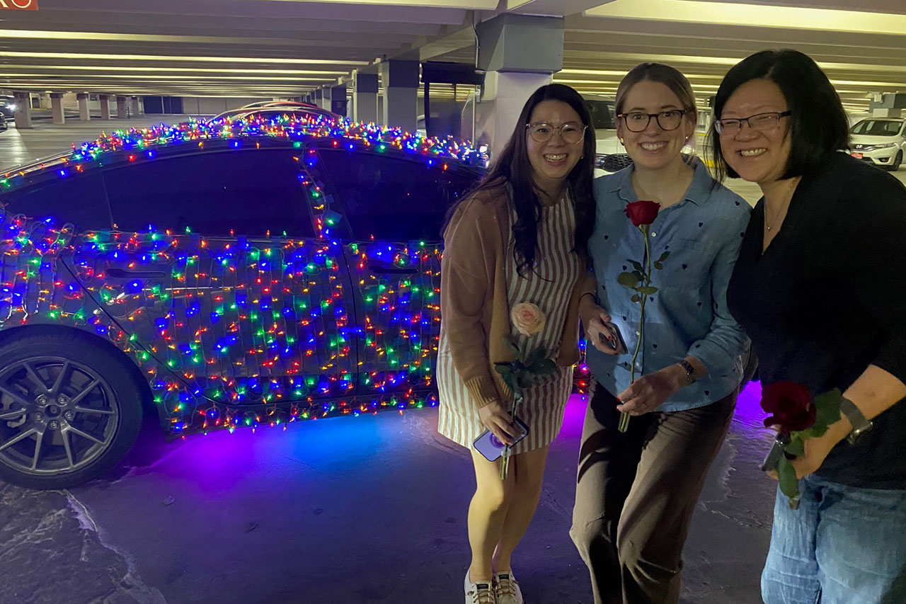 Three women in front of a car that is covered in Christmas lights