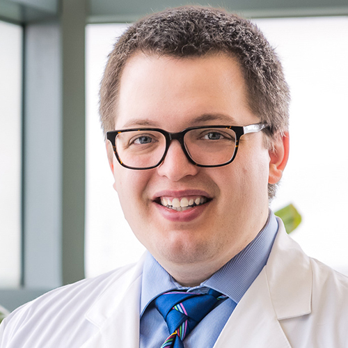 Dr. Crisologo, a smiling man with brown hair and glasses, wearing a blue shirt and tie under his lab coat.