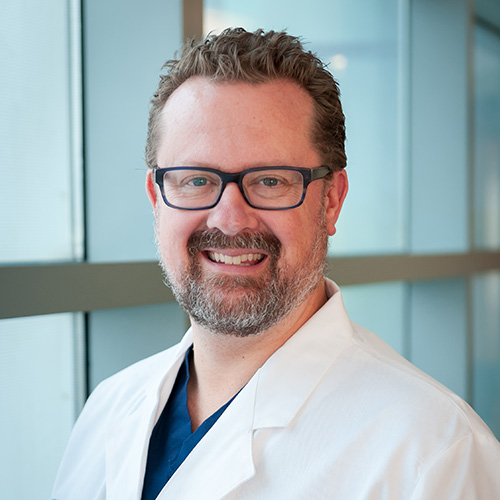 Dr. Sammer, smiling man with curly brown hair, beard and mustache, wearing a lab coat and glasses.