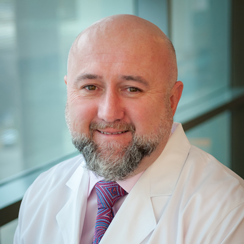 Dr. Hoxworth, a smiling man with a shaved head, gray mustache and beard, wearing a white lab coat.