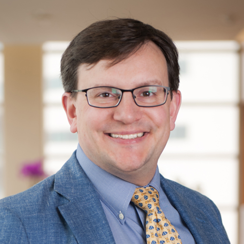 Dr. Seaward, a smiling man with dark hair and glasses, wearing a lab coat over a blue shirt and yellow patterned tie.