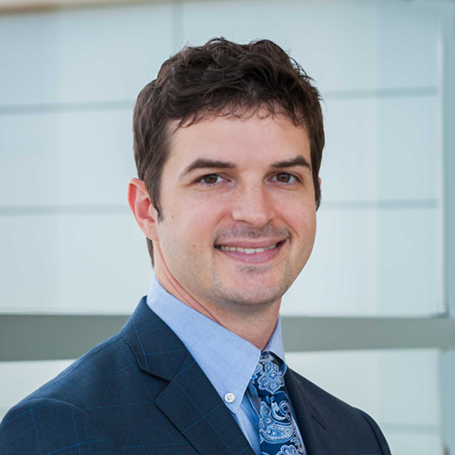 Dr. Lies, a smiling man with dark, curly hair wearing a dark suit.