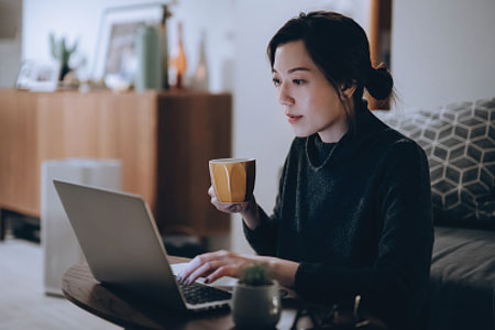 A woman holding a mug, scrolling on laptop while sitting on the couch